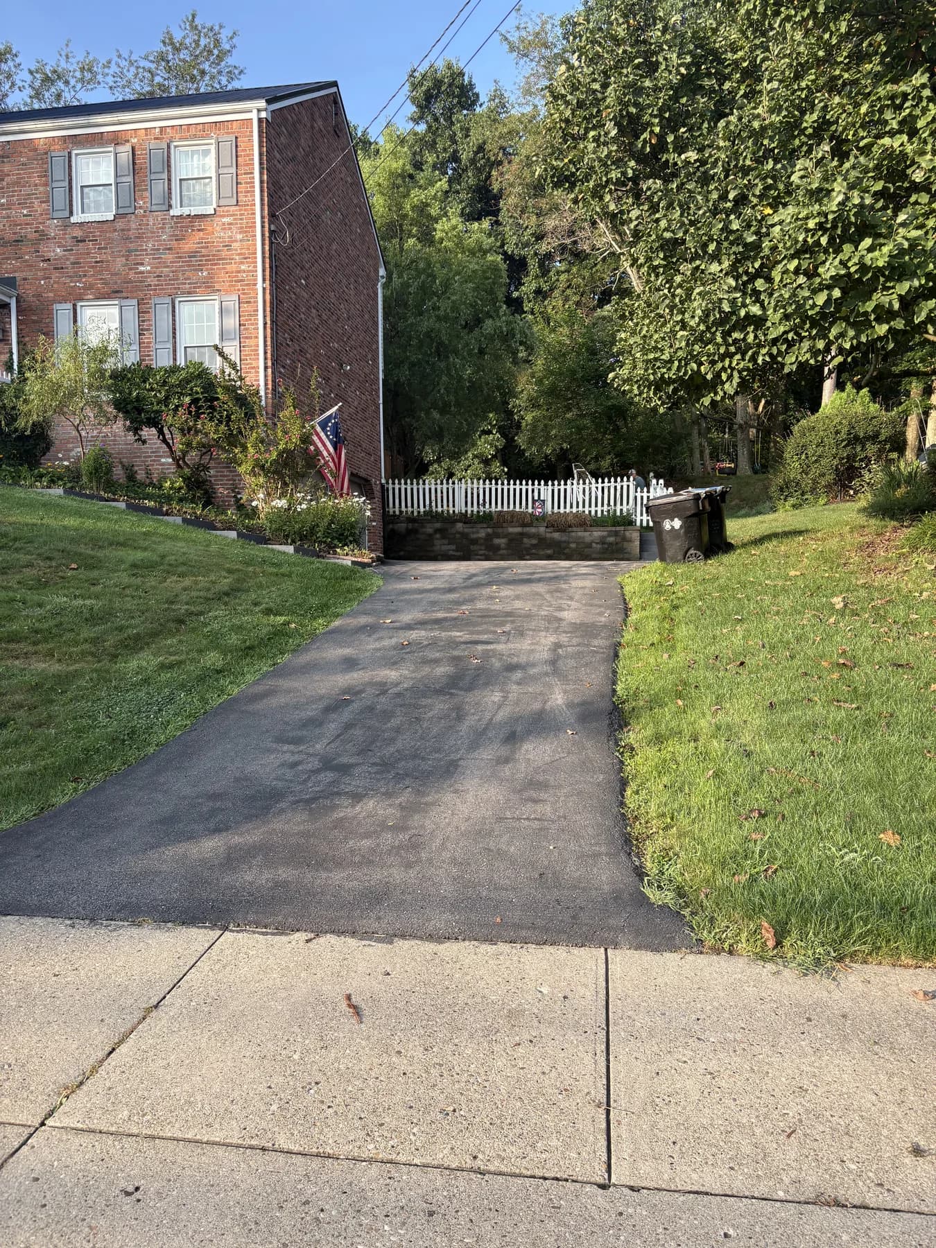 Faded gray asphalt driveway before sealcoating at a red-brick home with a white picket fence